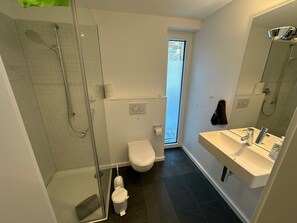 Et moderne badeværelse med brusekabine i glas, toilet, vask med spejl og et vindue, der giver naturligt lys.