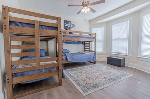 A room with two bunk beds, a ceiling fan, a rug, and a window with blinds. - Uptown Cottage | Near Strand +Postoffice + UTMB (Galveston)