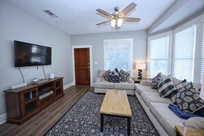 A living room with a flat-screen TV, wooden entertainment center, ceiling fan, and a sofa with patterned cushions. - Uptown Cottage | Near Strand +Postoffice + UTMB (Galveston)