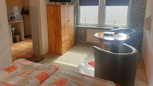 Een compacte kamer met kitchenette, televisie, raam met zonwering, klein rond tafeltje en een bank.
