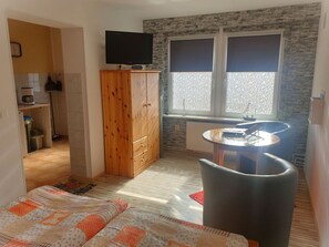 Ein kleines Zimmer mit Küchenzeile, Fernseher, Fenster mit Jalousien, einem kleinen runden Tisch und einer Couch.