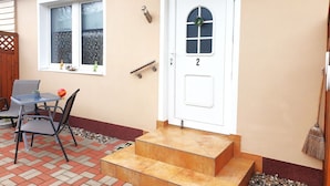 Een huis met een witte deur waarop het cijfer 2 staat, een klein portaal met een tafel en stoelen, en een raam met een krans.
