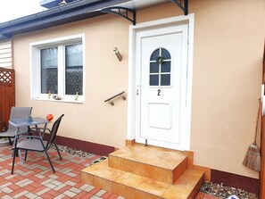 Ein Haus mit weißer Tür, beschriftet mit der Nummer 2, einem kleinen Vorbau mit Tisch und Stühlen sowie einem Fenster mit einem Kranz.