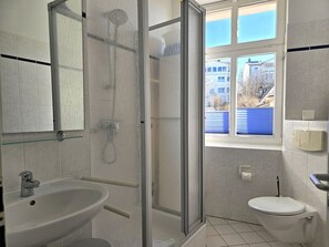 Ein Badezimmer mit Duschkabine aus Glas, Waschbecken, Toilette und Fenster mit Blick auf Gebäude.