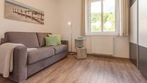 Ein Wohnzimmer mit einer Couch, einer Stehlampe, einem Fenster mit Vorhängen, einer Heizung und einem Bild an der Wand.