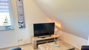 Ein modernes Wohnzimmer mit einem Flachbildfernseher auf einem Holzboden, zwei schwarzen Ledersesseln, einem Couchtisch mit Dekoration und einem Modell eines Segelbootes auf dem Fensterbrett.