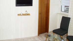 Ein Zimmer mit einem an der Wand befestigten Fernseher, einer Holztür, einem Glastisch und einer Fernbedienung sowie einem schwarzen Stuhl.