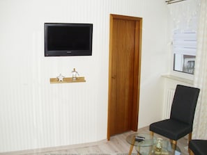 Ein Zimmer mit einem an der Wand befestigten Fernseher, einer Holztür, einem Glastisch und einer Fernbedienung sowie einem schwarzen Stuhl.