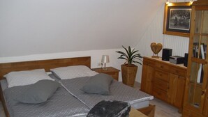Ein ordentlich eingerichtetes Schlafzimmer mit Bett, Nachttisch, Lampe, Blumentopf, Holzkommode und einem gerahmten Bild an der Wand.