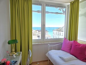 A room with a bed, a window with curtains, a radiator, and a table with a lamp and decorative items. - Villa Seeblick App. 308 - with magnificent sea view - Villa Seeblick, App. 308 - (Sassnitz auf Rügen)