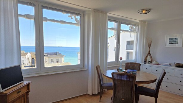 Ett rum med ett matbord, stolar, en tv-apparat på ett träskåp och stora fönster med utsikt över havet.