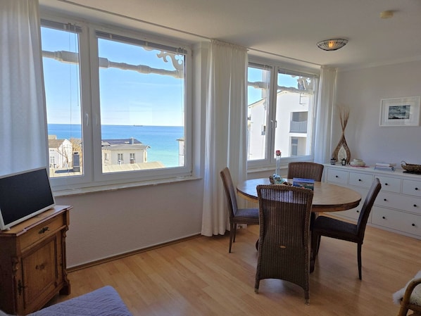 A room with a dining table, chairs, a TV on a wooden cabinet, and large windows with a view of the sea. - Villa Seeblick App. 308 - with magnificent sea view - Villa Seeblick, App. 308 - (Sassnitz auf Rügen)