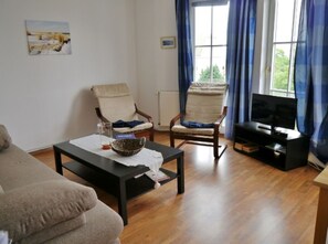 Ein Wohnzimmer mit einer Couch, zwei Sesseln, einem Couchtisch, einem Fernseher und einem Fenster mit Vorhängen.