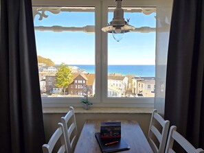 Ein Zimmer mit Tisch und Stühlen, ein Fenster mit Blick auf Gebäude und das Meer sowie einer Deckenleuchte.