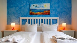 A double bed with white linens, two bedside tables with lamps, and a painting of a beach scene on the wall. - Vacation apartment Fraenzi - with wonderful sea view - Ferienappartement Fraenzi (Sassnitz auf Rügen)