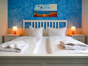 A double bed with white linens, two bedside tables with lamps, and a painting of a beach scene on the wall. - Vacation apartment Fraenzi - with wonderful sea view - Ferienappartement Fraenzi (Sassnitz auf Rügen)