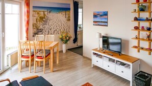 A modern living room with a beach mural, a dining area, a TV, and a bookshelf. - Vacation apartment Fraenzi - with wonderful sea view - Ferienappartement Fraenzi (Sassnitz auf Rügen)