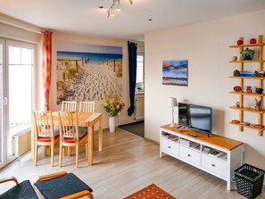 A modern living room with a beach mural, a dining area, a TV, and a bookshelf. - Vacation apartment Fraenzi - with wonderful sea view - Ferienappartement Fraenzi (Sassnitz auf Rügen)