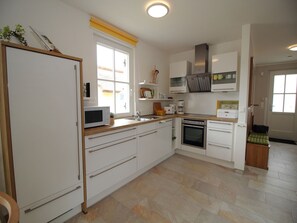 A modern kitchen with white cabinetry, a built-in oven, microwave, and a large window with a yellow blind. - Reethaus Seeschwalbe - close to the beach - Reethaus Seeschwalbe close to the beach (Glowe auf Rügen)