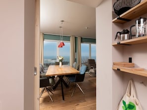 Ein moderner Essbereich mit Holztisch, Stühlen, einer hängenden roten Pendelleuchte und Meerblick durch große Fenster.