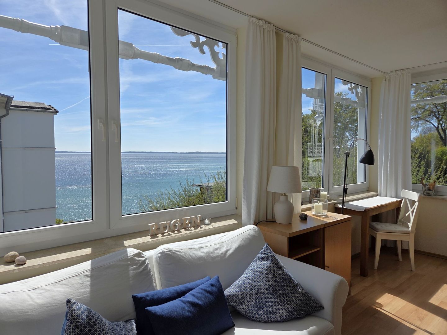 A room with a large window overlooking a body of water, a white sofa with blue cushions, a wooden desk, and a chair.