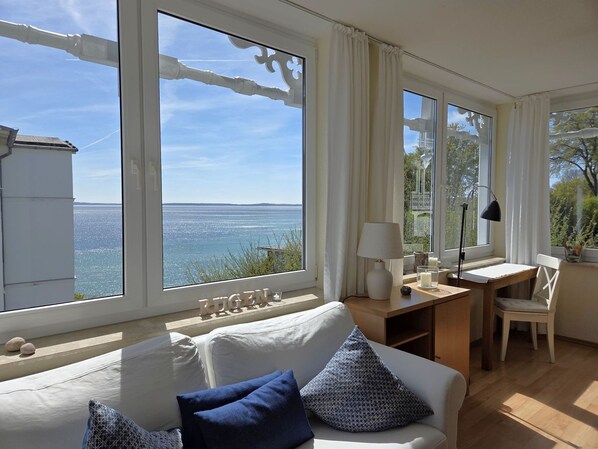 Ein Zimmer mit einem großen Fenster mit Blick auf ein Gewässer, ein weißes Sofa mit blauen Kissen, ein Holztisch und ein Stuhl.