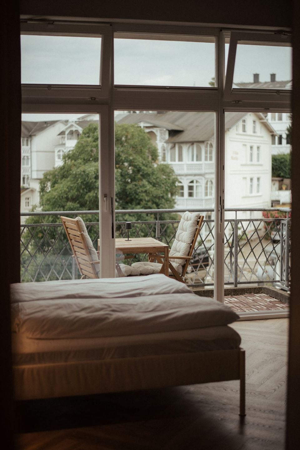 Un dormitorio con una cama grande, una mesita pequeña y dos sillas en un balcón con vistas a edificios.