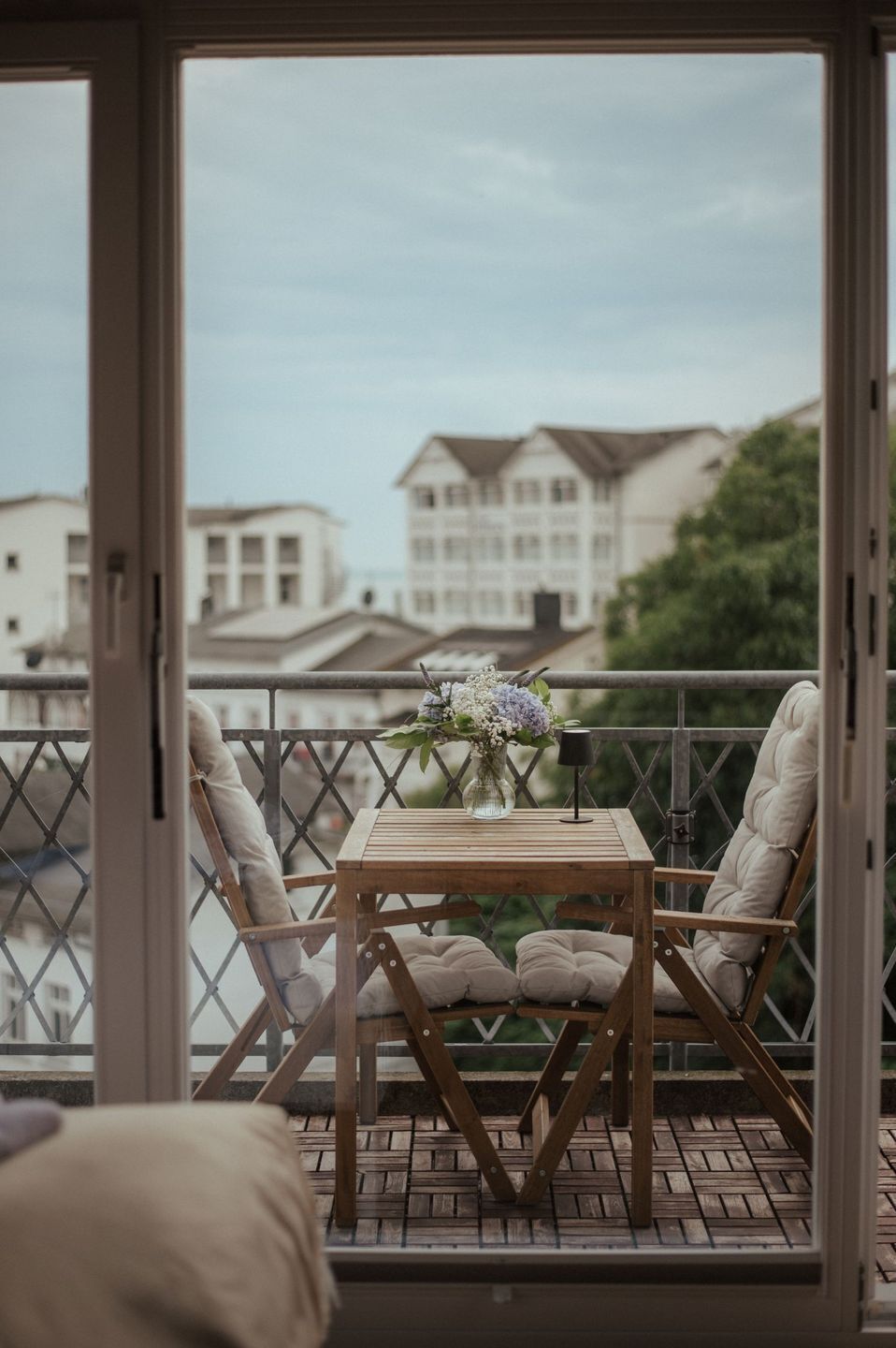 A balcony with a wooden table and two cushioned chairs, a vase with flowers, and a view of buildings in the background.