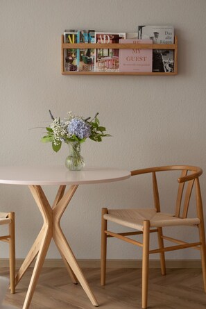 Ein runder Tisch mit Blumen in einer Vase, zwei Holzstühle und ein an der Wand befestigtes Regal mit Zeitschriften und Büchern.