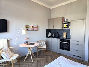 A compact room with a kitchenette, a small dining table, a chair, a bed, and a wall-mounted TV. - Alter Reichshof, Apartment 303 with Ocean View and Balcony - Ocean View & Balcony in Alt (Sassnitz auf Rügen)