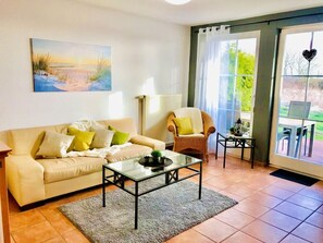 Ein Wohnzimmer mit einer Couch, einem Glastisch, einem Korbstuhl und Blick durch eine Glastür.