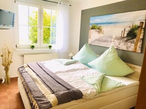 Ein Schlafzimmer mit Bett, einem Fenster mit Vorhängen, einem Heizkörper, einem an der Wand befestigten Fernseher und einem Bild mit Strandmotiv.