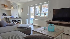 A modern living room with a sofa, a coffee table, a TV, and a window with a view of buildings. - Seemeile 1 - Villa Monika - Villa Monika, App. Seemeile 1 with terrace (Sassnitz auf Rügen)