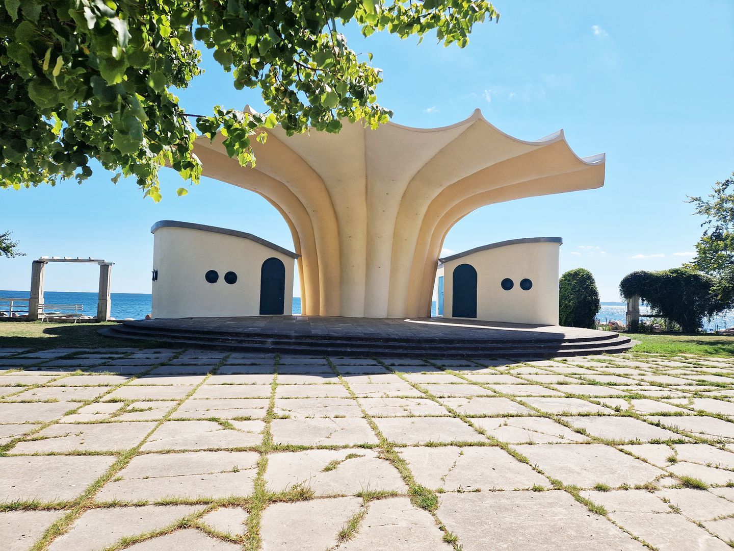 A symmetrical architectural structure with a central archway and two smaller arches on either side, set on a paved area with grass patches.