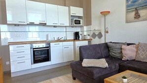 A compact living space with a kitchenette, a sofa, a coffee table, and a painting of a beach scene. - Fürstenhof, App.104 - First row by the sea - Fürstenhof, App. 104 - First row (Sassnitz auf Rügen)