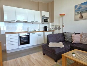 A compact living space with a kitchenette, a sofa, a coffee table, and a painting of a beach scene. - Fürstenhof, App.104 - First row by the sea - Fürstenhof, App. 104 - First row (Sassnitz auf Rügen)