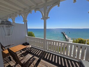 Ein überdachter Außenbereich mit Holztisch und -stühlen, einem weißen Geländer und Blick auf das Meer und einen Steg.