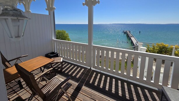 A covered outdoor area with a wooden table and chairs, a white railing, and a view of the ocean and a pier. - Fürstenhof, App.104 - First row by the sea - Fürstenhof, App. 104 - First row (Sassnitz auf Rügen)