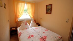 Una camera da letto con un letto, due cuscini, un comodino, una finestra con tende e un quadro appeso al muro, oltre a una presa elettrica.