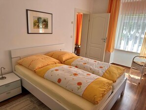 Ein Doppelbett mit floralem Bettbezug, ein Nachttisch, ein Stuhl, eine Tür und ein Fenster mit Vorhängen.