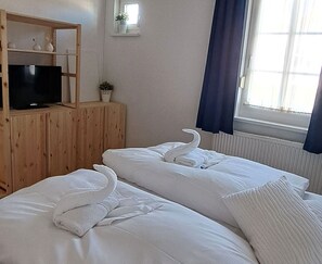 Ein Hotelzimmer mit zwei Betten, einem Fernseher auf einer Holzkommode, einer kleinen Pflanze und einem Fenster mit Vorhängen.
