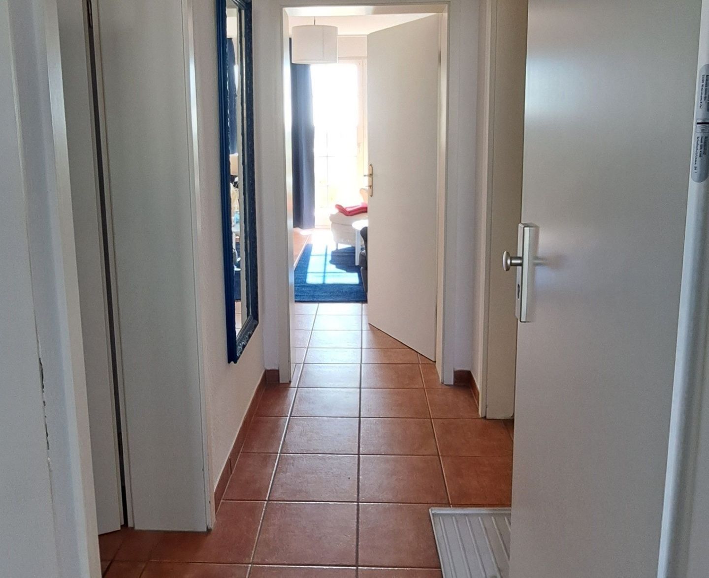 Ein Flur mit Fliesenboden, einer Tür mit silbernem Griff und einem Spiegel an der linken Wand.