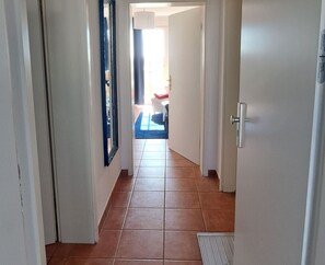 Ein Flur mit Fliesenboden, einer Tür mit silbernem Griff und einem Spiegel an der linken Wand.