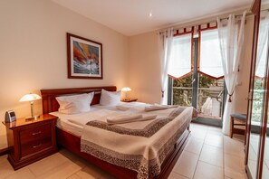A hotel room with a bed, bedside lamps, a nightstand, a wooden headboard, a painting, and a balcony with curtains. - Villa Friede-Marie - Penthouse Sophie - Villa Friede-Marie, Penthouse Sophie - (Sassnitz auf Rügen)