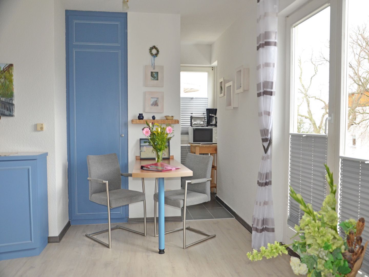 Ein Zimmer mit blauer Tür, einem kleinen Tisch mit zwei Stühlen, einer Vase mit Blumen und einem Fenster mit Jalousien.