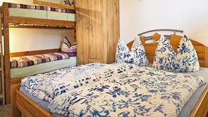 Een stapelbed met een onderbed, een houten garderobe, een bed met een bloemenprintend beddegoed, en een plank met decoratieve objecten.