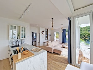 Eine moderne Küche mit Marmorarbeitsplatte, Holzschneidebrett und Kochutensilien. Im Hintergrund ist ein Wohnbereich mit Sofa und Esstisch zu sehen.
