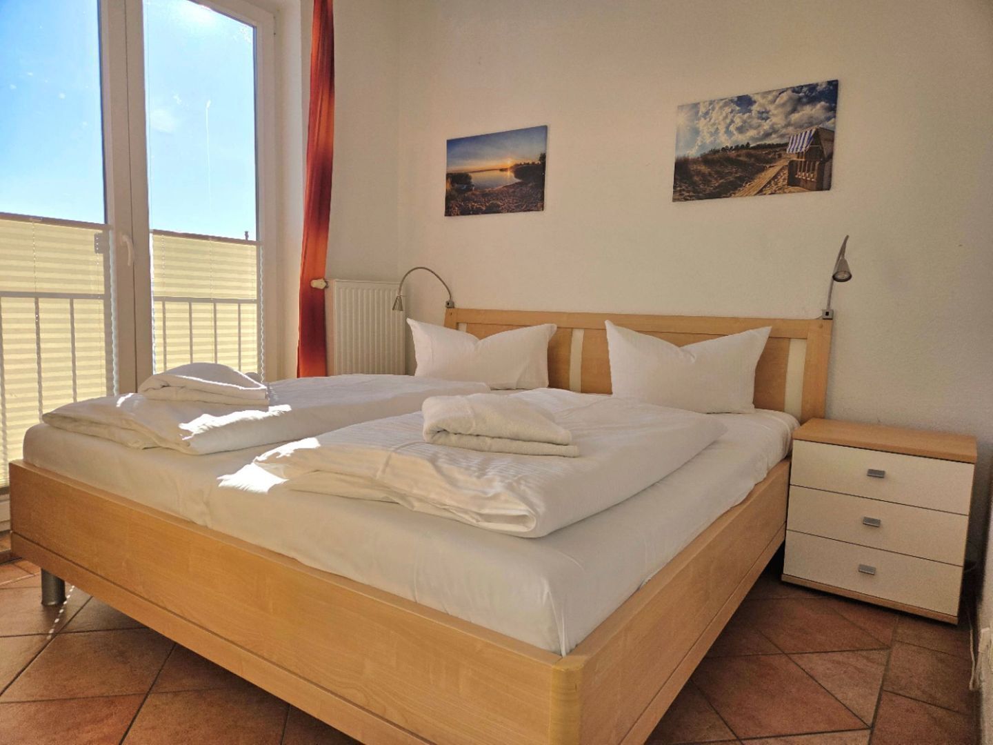 Ein Doppelbett mit weißer Bettwäsche, ein hölzerner Kopfteil, ein Nachttisch mit Schubladen sowie zwei an der Wand befestigte Bilder.