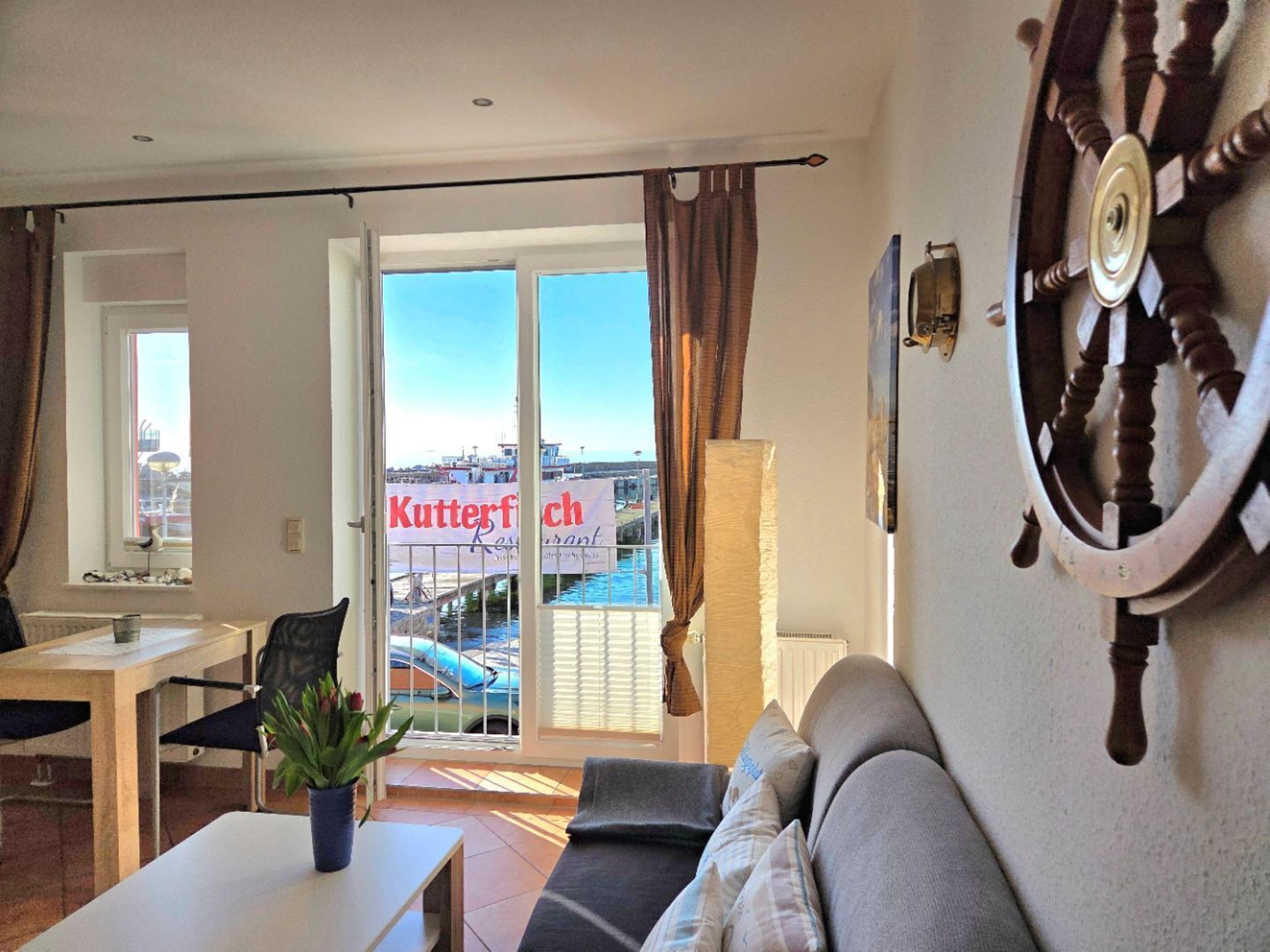 Ein Wohnzimmer mit einer Couch, einem Esstisch, einer Topfpflanze und Blick auf das Meer. Ein Schild zeigt 'Kutterfisch'.