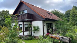 Ein zweistöckiges Haus mit braunem Dach und weißen Wänden, umgeben von üppiger Vegetation.